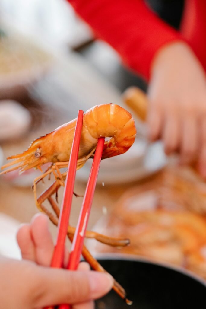 pexels photo 7363731 7363731 Close-up of a person using red chopsticks to pick up freshly cooked shrimp at a dining table.